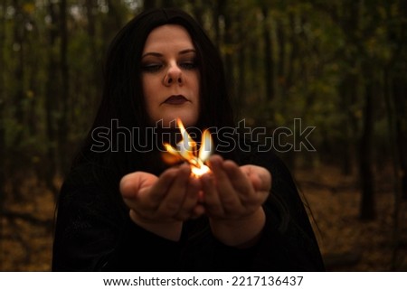 Black-haired witch holds fire in her hands in the forest.