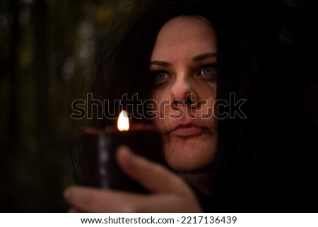 Black-haired witch holding candle in her hands in the forest.