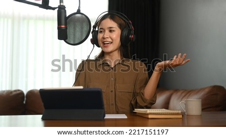 Smiling young woman wearing headphone and talking through microphone recording podcast in small home studio