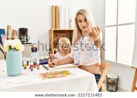 Caucasian family of mother and daughter painting at art studio pointing with finger to the camera and to you, confident gesture looking serious 