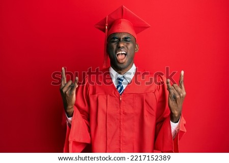 Young african american man wearing graduation cap and ceremony robe shouting with crazy expression doing rock symbol with hands up. music star. heavy concept. 
