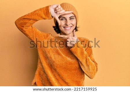 Young brunette woman wearing wool hat and winter sweater smiling making frame with hands and fingers with happy face. creativity and photography concept.  Royalty-Free Stock Photo #2217159925