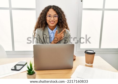 Beautiful hispanic business woman sitting on desk at office working with laptop cheerful with a smile of face pointing with hand and finger up to the side with happy and natural expression on face 