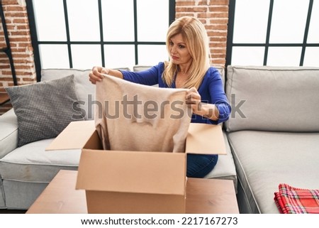 Middle age blonde woman sitting on the sofa opening a box with sweater skeptic and nervous, frowning upset because of problem. negative person. 