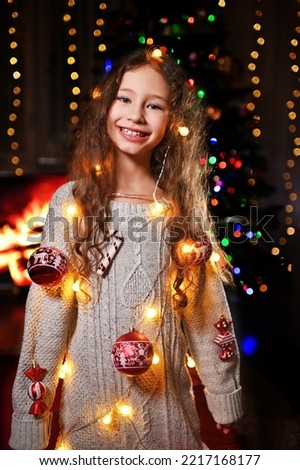 a cute girl with curly hair in a knitted sweater with Christmas toys smiles and depicts a Christmas tree against the background of a fireplace and Christmas lights.