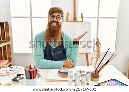 Redhead man with long beard painting clay bowl at art studio smiling cheerful presenting and pointing with palm of hand looking at the camera. 