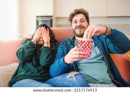 Scared millennial newlywed couple watching horror movie on tv, frightened young woman covering eyes and man laughing and eating popcorn during a thrilling scary film moment sitting on sofa at home Royalty-Free Stock Photo #2217176913
