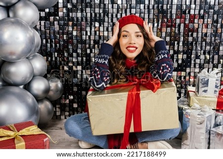The woman rejoices and hides her face with her hands. Beautiful brunette in a Christmas hat, scarf and sweater on a silver background.