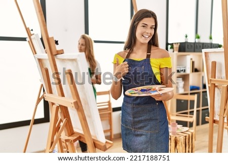 Young hispanic woman at art classroom smiling happy and positive, thumb up doing excellent and approval sign 