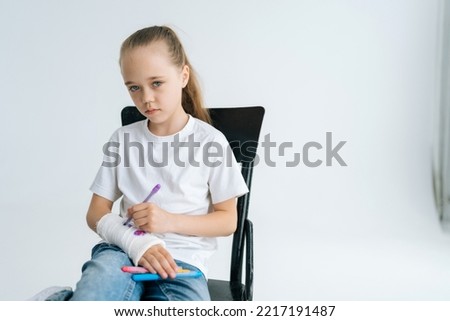 Portrait of serious little girl drawing cute image with colorful marker on broken arm wrapped in white plaster bandage looking at camera. Concept of child insurance and healthcare.