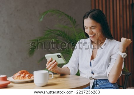 Young latin woman wear white shirt play app video games on mobile cell phone gadget smartphone do winner gesture sit at table in coffee shop cafe restaurant indoors. Freelance office business concept Royalty-Free Stock Photo #2217236283