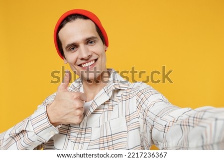 Close up young smiling happy cheerful caucasian man 20s wearing white casual shirt orange hat doing selfie shot pov on mobile phone show thumb up gesture isolated on plain yellow background studio.