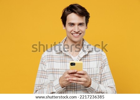 Young smiling happy cheerful european man 20s in white casual shirt hold in hand use mobile cell phone browsing internet isolated on plain yellow background studio portrait. People lifestyle concept
