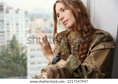 Woman in camouflage uniform with serious expression sits on windowsill