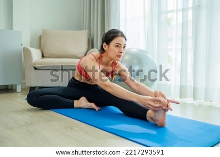 Young beautiful asian girl is doing yoga at home. She is stretching on a blue yoga mat. She is wearing fitness clothing. She is watching her instructor on the tablet.