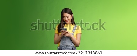 Outgoing cute asian brunette woman scroll online shop, purchase summer tour internet, hold smartphone, look amused telephone screen, use app, edit photos post social media, stand green background.