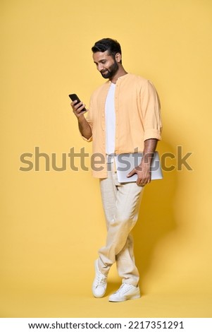 Smiling indian man user, student or employee standing isolated on yellow background holding laptop using mobile phone advertising digital products for work and learning. Full length vertical shot.