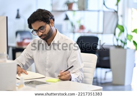 Busy indian professional business man, corporate employee, eastern businessman executive worker working at office desk planning agenda writing on sticky notes managing tasks sitting at workplace.