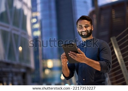 Smiling happy young eastern indian business man professional manager standing outdoor on street holding using digital tablet online fintech in night city with urban lights looking at camera, portrait. Royalty-Free Stock Photo #2217353227