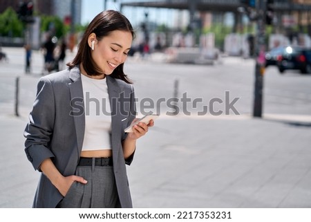 Young professional Asian business woman wearing suit using wireless earphones holding smartphone talking on mobile phone having chat on cellphone walking on urban city street. Royalty-Free Stock Photo #2217353231
