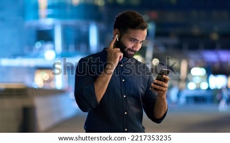Smiling bearded ethnic indian man wearing earbud holding cell phone at night. Eastern businessman in earphone using smartphone listening music in app tech on cellphone watching videos online. Royalty-Free Stock Photo #2217353235