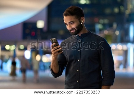 Rich successful indian business man professional entrepreneur wearing earbud standing outdoors on night city street holding cell phone using smartphone watching video or having mobile chat call. Royalty-Free Stock Photo #2217353239