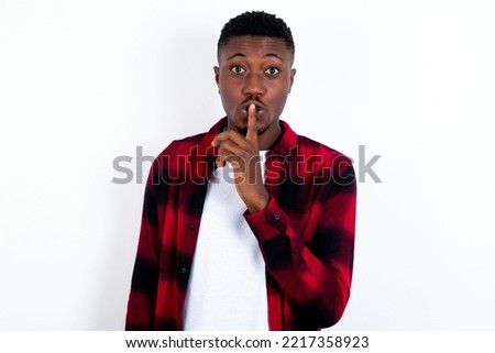 Surprised young handsome man wearing red plaid shirt over blue background makes silence gesture, keeps finger over lips and looks mysterious at camera