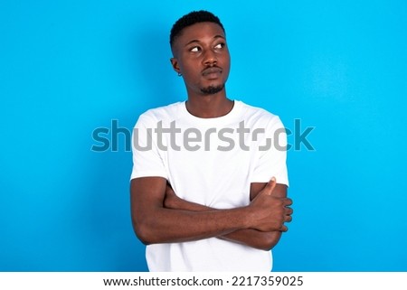 Pleased young handsome man wearing white T-shirt over blue background keeps hands crossed over chest looks happily aside