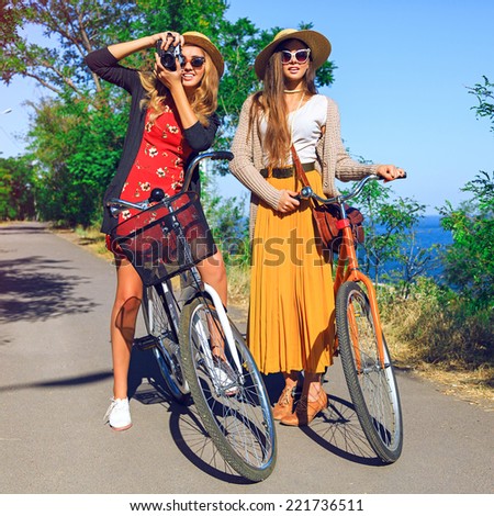Two pretty young playful girls, walking near sea side with retro bikes, best friends having fun taking pictures of each other, wearing stylish vintage clothes sunglasses and hats.