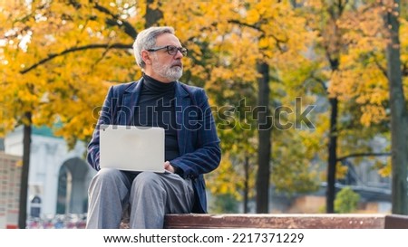 Modern stylish bearded grey-haired mature man entrepreneur sitting in the park with fall foliage and working on his laptop. High quality photo