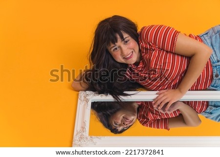 Young, happy, caucasian girl with long brown hair laying near close to the mirror on the floor. Girls smiling reflection in the mirror, isolated orange background. High quality photo