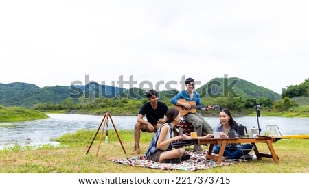 Group of Asian man and woman friends singing and playing guitar during having breakfast by the lake together. People enjoy and fun outdoor lifestyle travel nature camping on summer holiday vacation. Royalty-Free Stock Photo #2217373715
