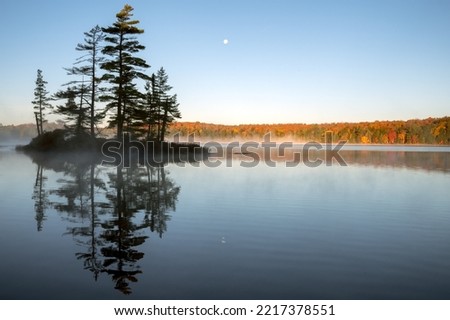 An island reflects on a calm lake with an autumn colored, tree lined shoreline and the moon in the background. Islet Lake, Algonquin Provincial Park, Ontario, Canada. Royalty-Free Stock Photo #2217378551