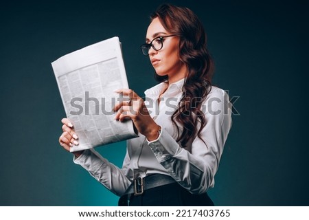 Beautiful young business woman in glasses holds newspaper in her hands and reads carefully. Study of news edition. Caucasian woman reads press on dark background. Studio portrait. Bottom view of hero.