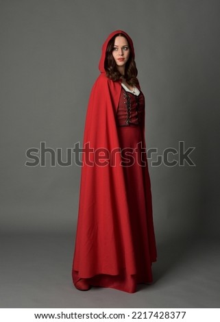 Full length portrait of woman wearing red medieval fantasy costume, flowing hooded cloak. Standing pose inside profile, gestural hand poses, walking away from camera isolated on grey studio background Royalty-Free Stock Photo #2217428377