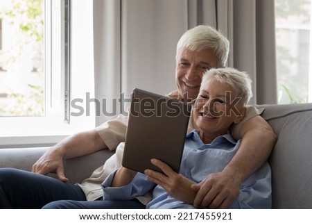 Happy affectionate middle aged old 60s retired family couple using digital computer tablet, resting on couch, enjoying watching funny photo video, choosing goods shopping online or web surfing info.