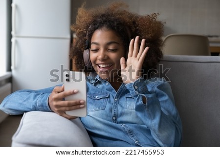 Attractive African teenager girl in wireless headphones greets friend start video call talk using smartphone seated on sofa at home. Distancing communication using modern tech, virtual meeting event Royalty-Free Stock Photo #2217455953