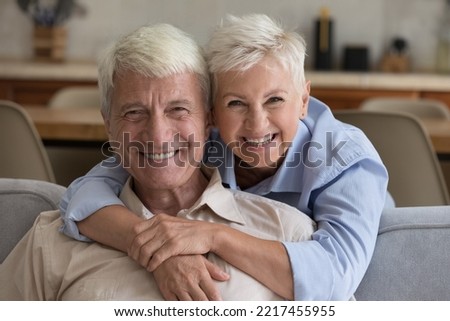 Grey-haired grandparents pose at home. Loving wife hugs husband seated on sofa staring at camera, spend time together. Happy long-lasting marriage, harmonic relations between spouses, respect and bond Royalty-Free Stock Photo #2217455955