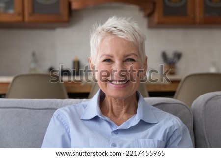 Head shot portrait older blonde woman resting on sofa staring at camera feels happy, having wide toothy smile and attractive appearance, enjoy retired carefree life at home alone looking optimistic