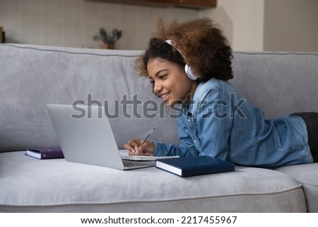 African teenager girl, high school student lying on sofa use laptop writes assignment in copybook, listens on-line tutor or audio course, e-learn use modern tech, participates in educational webinar Royalty-Free Stock Photo #2217455967