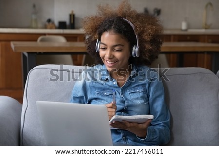 African student girl sit on sofa wear headphones takes notes, studying remotely use laptop and video call application. Gen Z and modern tech usage, distancing learning at home, tuition, online class Royalty-Free Stock Photo #2217456011