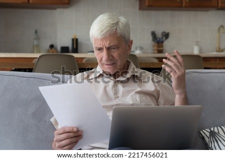 Older man sit on sofa with laptop feels angry looks dissatisfied read unpleasant notification from bank, get paper notice, looks annoyed due to overspend, debt, taxes, bad news, financial difficulties Royalty-Free Stock Photo #2217456021