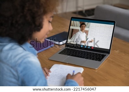 African teenager girl sit at desk with laptop, writes exercise, makes assignment, improve skills use modern tech, e-learning with online Indian teacher explain English languages tenses. Webinar event Royalty-Free Stock Photo #2217456047