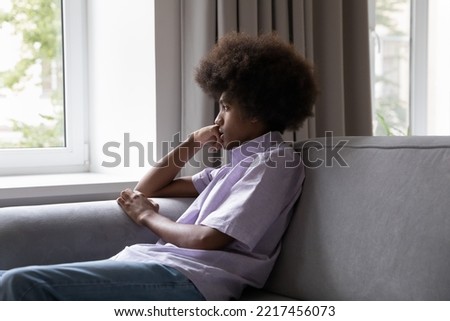 Sad African insecure teenage guy outcast sit on sofa staring through window, looks pensive thinks over life concerns or unrequited love, feels boredom at home alone on weekend. Teen worries, problems Royalty-Free Stock Photo #2217456073