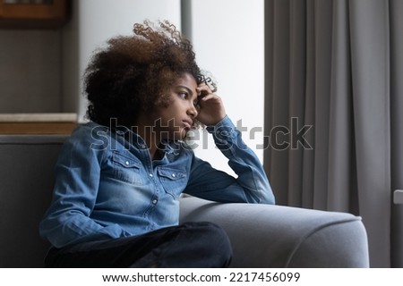 African teenage girl sits on sofa at home lost in thoughts, staring aside, thinking, looks upset experiences first unrequited love, having personal problems in school feels insecure. Teenage concerns Royalty-Free Stock Photo #2217456099