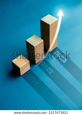 Shining rise up arrow on wooden cube blocks, bar graph chart steps on blue background with year numbers, 2023, 2022, 2021, vertical style. Business growth and development to success concepts. Royalty-Free Stock Photo #2217471821