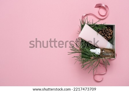 Beauty box of seasonal winter gifts on pink background. Cosmetic tube, serum and green fir branches with bump in cardboard packaging box with pink ribbon