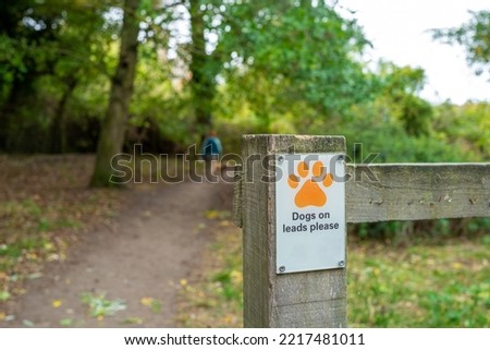Shallow focus of a laminated Dogs On Lead sign attached to a wooden fence at the entrance to a nature reserve. A woman and her dog can be seen.