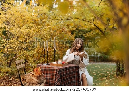Girl with a cup of coffee in her hands sits at a table in the autumn garden. Picnic in nature in autumn
