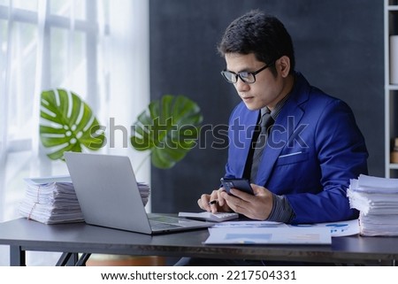 Young financial market analyst wearing glasses works in the office with his laptop while sitting at a wooden table. Asian businessman analyzing documents in hand with document graph